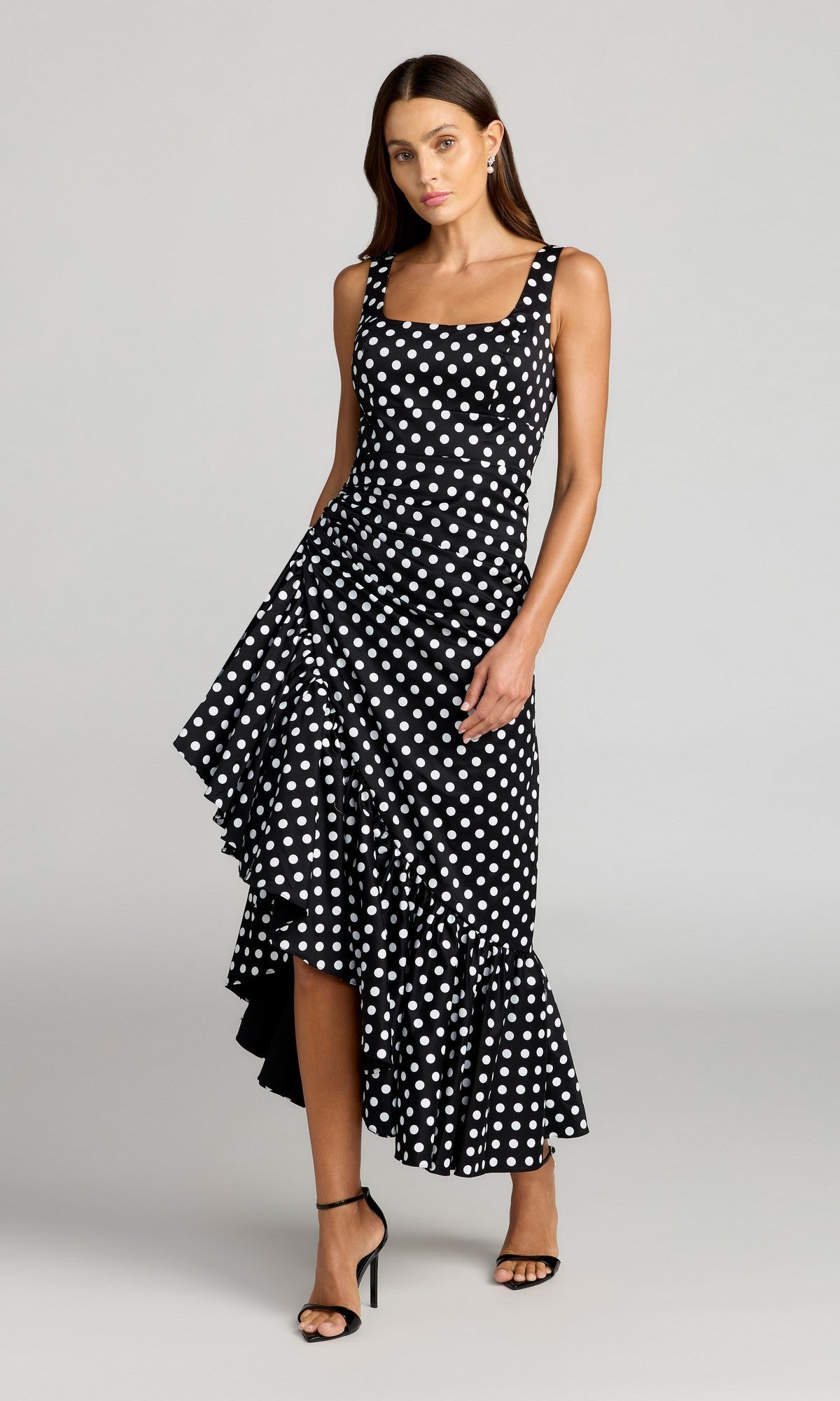 A woman wearing the Mac Duggal 27780 Midi Party Dress in Black Ivory––a sleeveless cocktail dress with white polka dots, a ruffled asymmetrical hem, and paired with black heeled sandals––stands against a plain light background. This elegant dress is designed to beautifully flatter the silhouette for any special occasion.